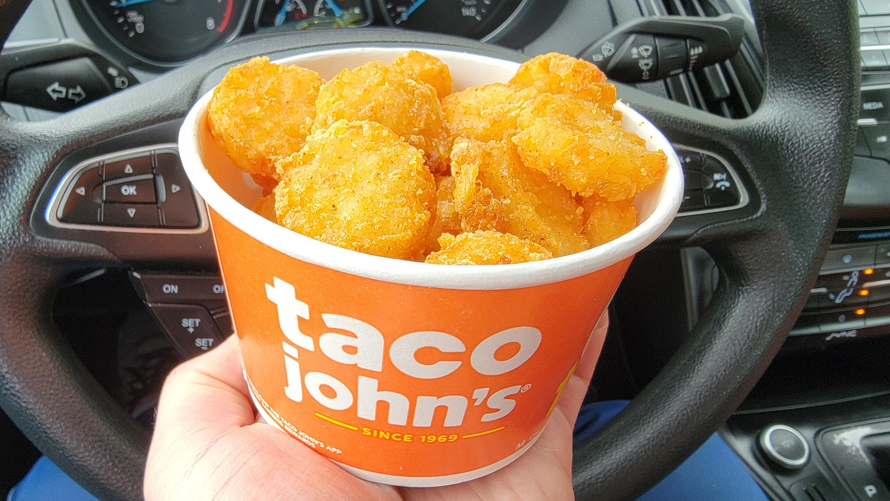 Cardboard cup of Taco John's Potato Olés held in front of a car steering wheel