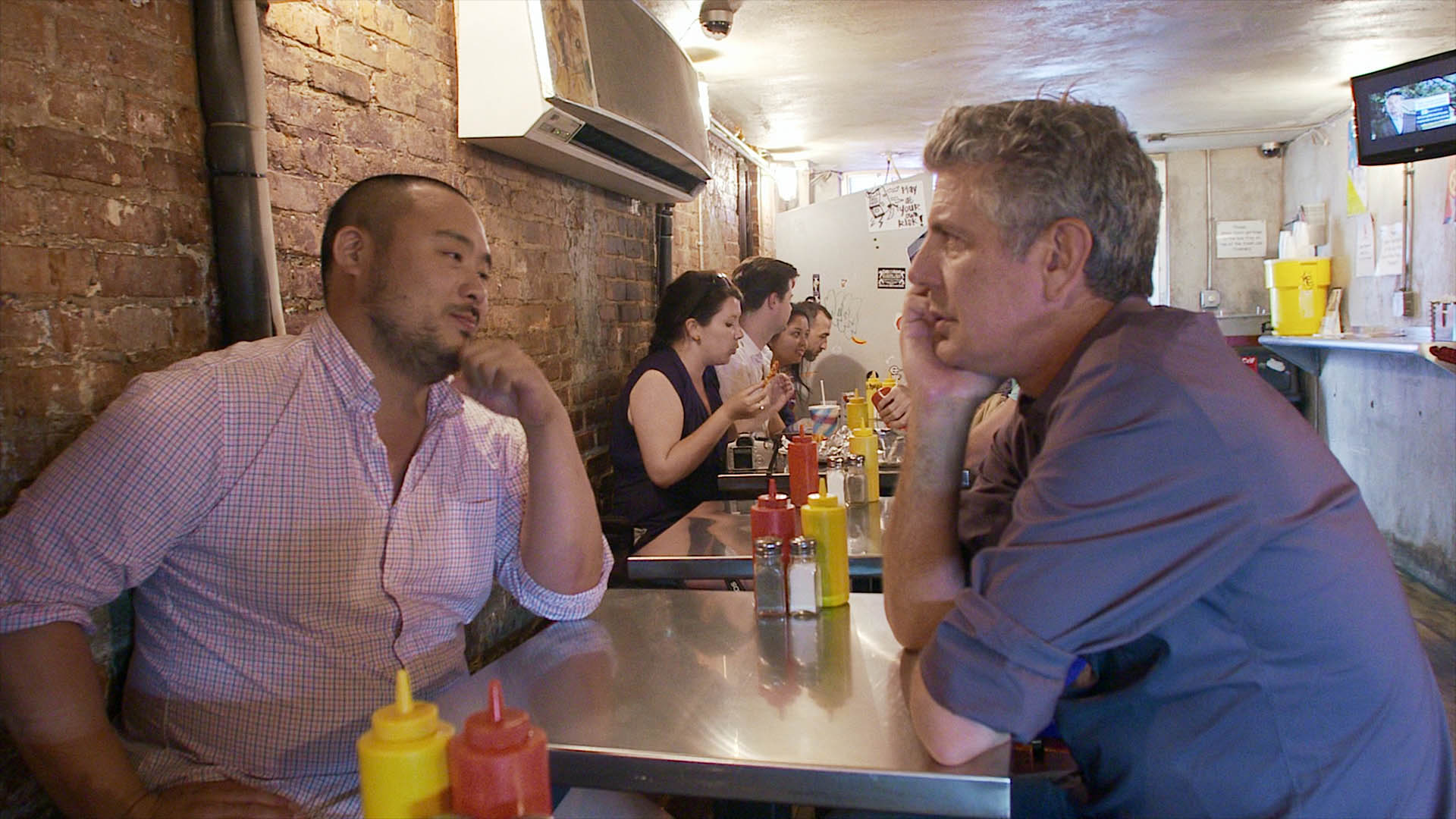 David Chang and Anthony Bourdain sitting at a restaurant table talking
