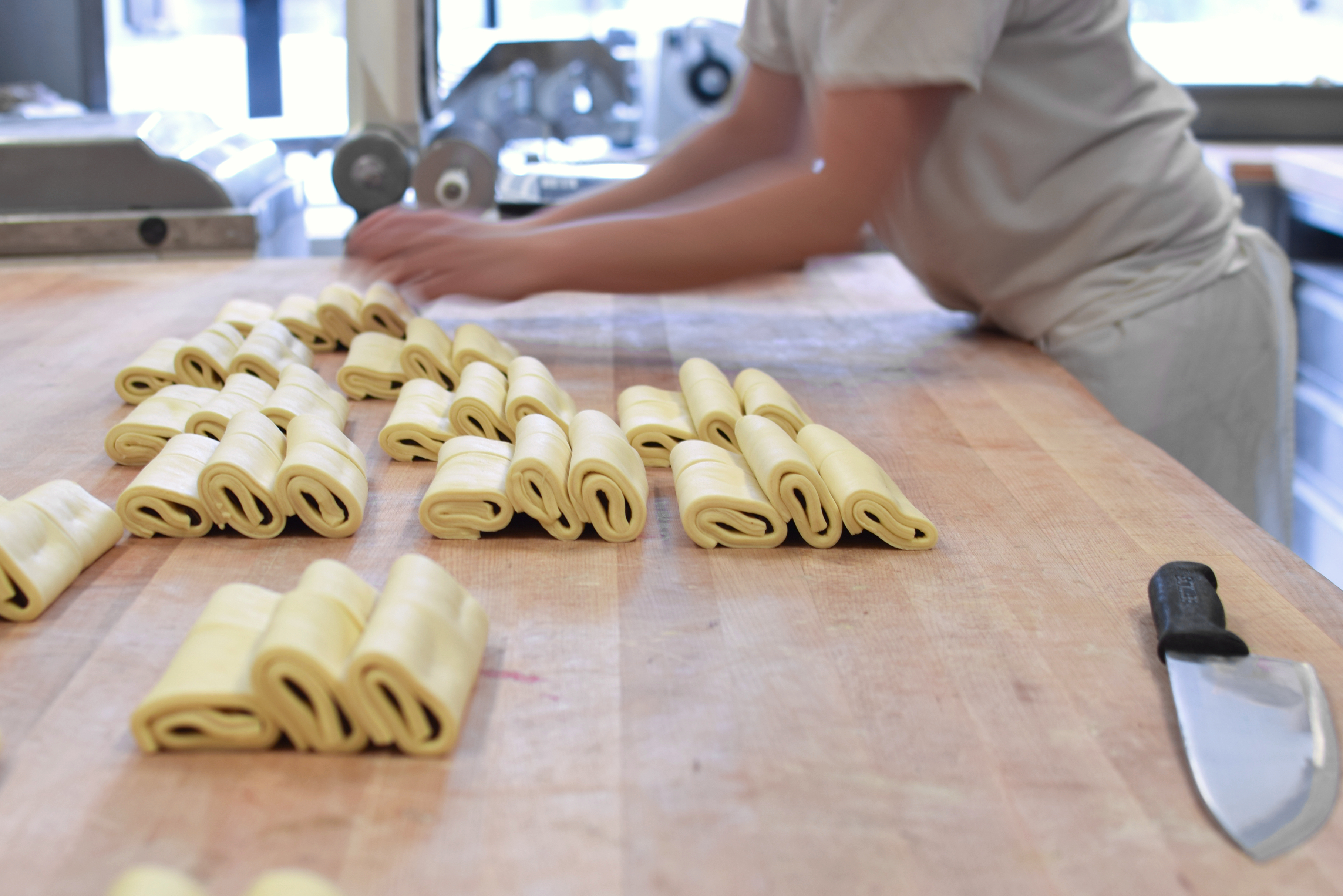 On a wooden work surface, chocolatines lie stacked up while a bakery employee folds them and stacks them.