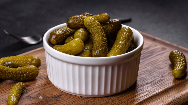 A ramekin of cornichons on wooden cutting board