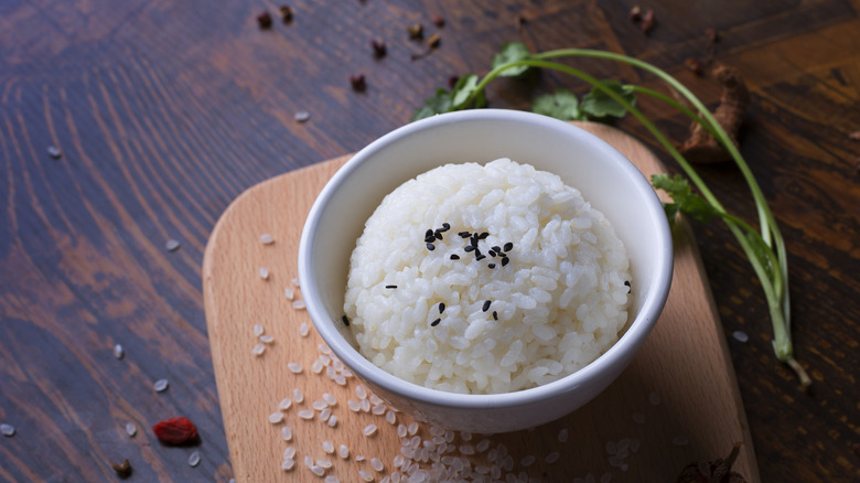 White bowl of cooked rice on cutting board next to herbs