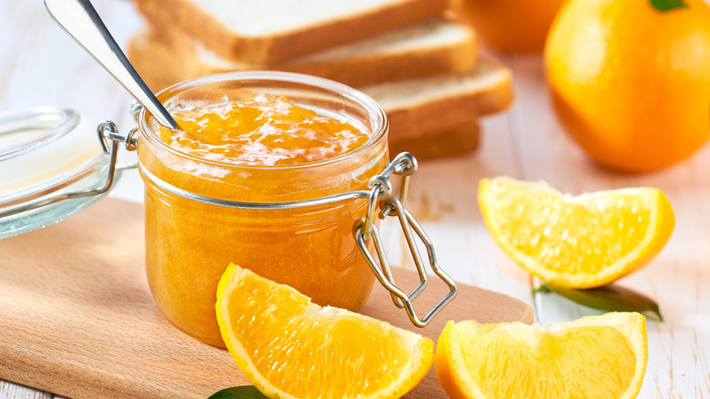 An open jar of orange marmalade with a spoon inside surrounded by orange slices