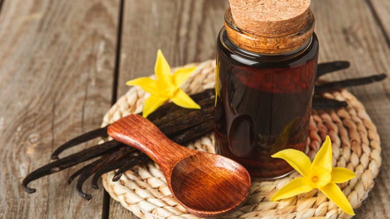 Vanilla extract in a glass bottle next to a wooden spoon, vanilla beans, and flowers