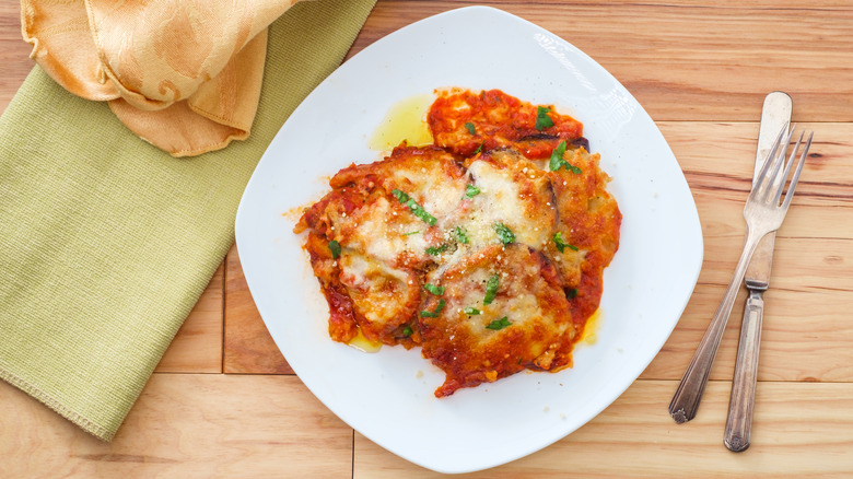 Eggplant Parm on white plate by silverware and cloth napkins