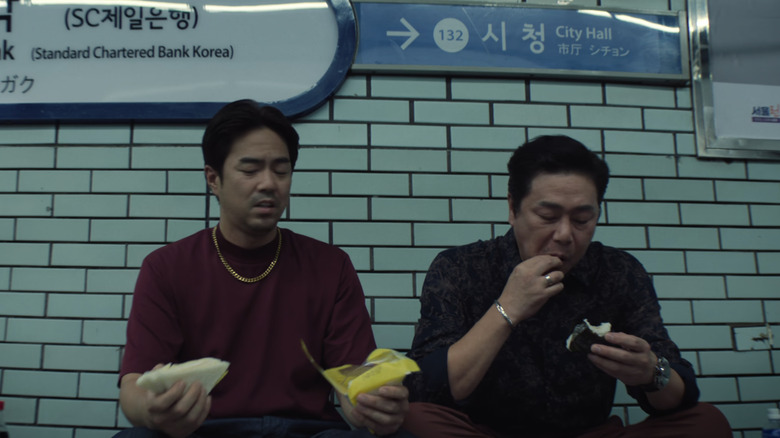 The two men react to and eat their food in the train station