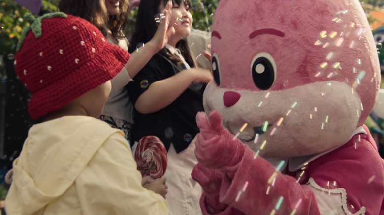 A bunny mascot hands a pink lollipop to a little girl in a strawberry hat