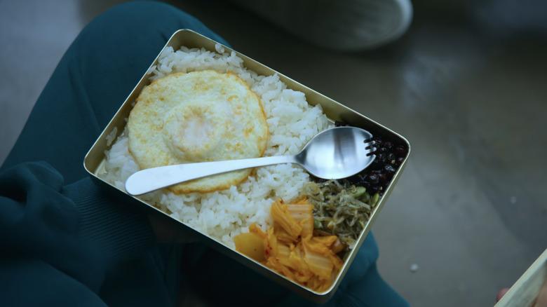 Silver rectangular lunch box with rice, kimchi, black beans, and a fried egg with a spork on top