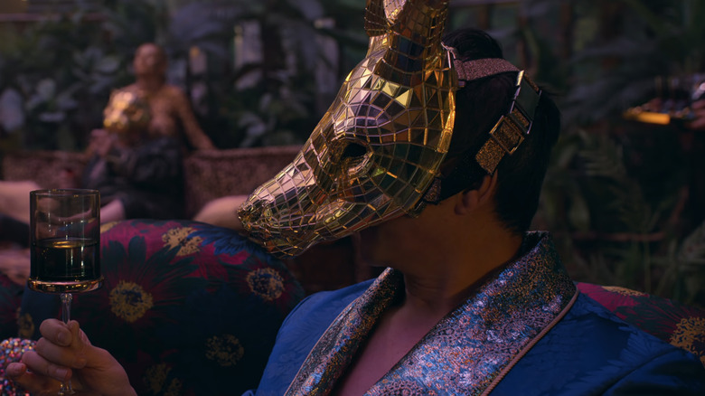 A VIP in a gold fox mask holds a cup of what appears to be scotch