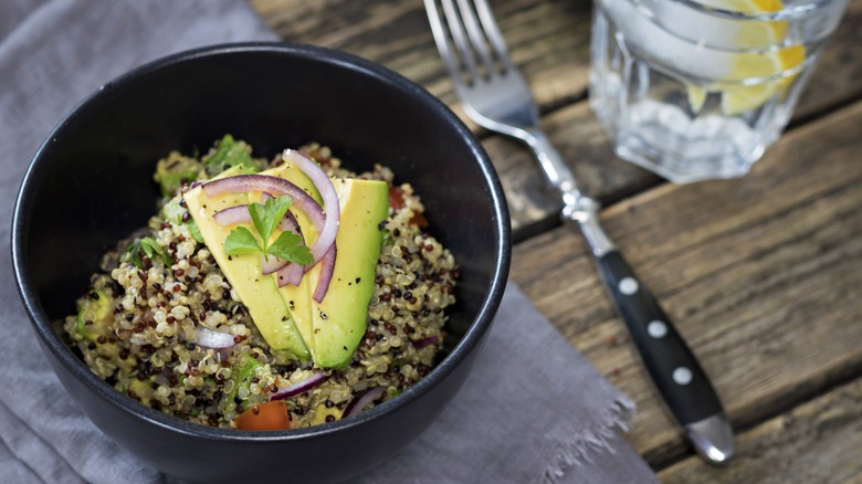 Bowl of quinoa topped with veggies and avocado beside a fork and glass of water