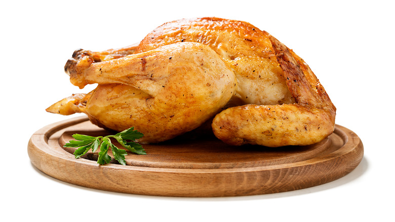 A whole roast chicken on a wooden platter and a sprig of parsley against a white backdrop