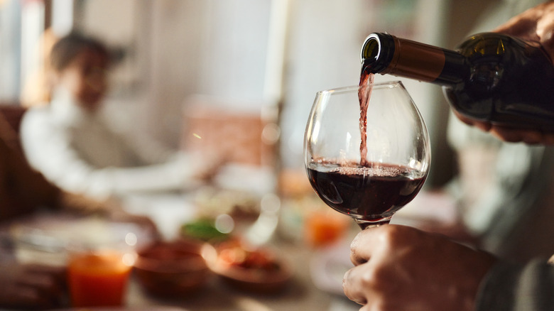 hands pouring glass of red wine with blurry person in background