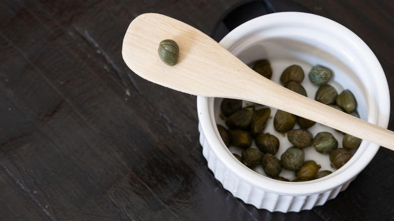 capers in a small white bowl with a small wooden paddle