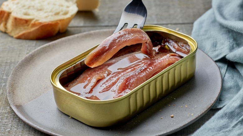 Fork lifting anchovy from open can on a plate