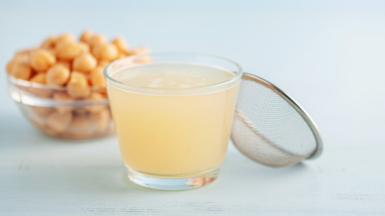 a small glass of aquafaba next to a strainer and bowl of chickpeas