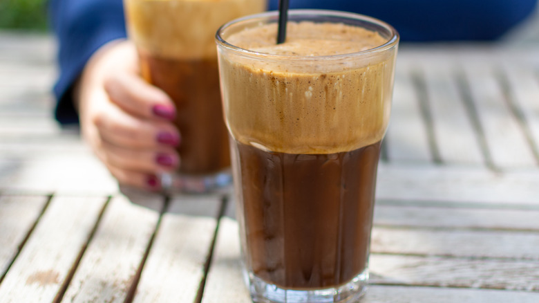 A frappe of the forefront, the coffee is darker at the bottom and foamy at the top, the glass cup has a black, plastic straw in it. On the background, a hand with a pink manicure and blue sleeve holds another identical drink.