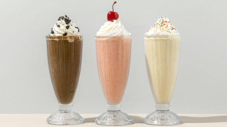 Three milkshakes in glass cups, chocolate with whipped cream and oreo crumbs, strawberry with whipped cream and a cherry, and vanilla with whipped cream and sprinkles on a white surface with a light grey background.