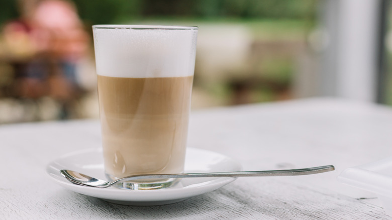 A glass cup of coffee with milk foam and a spoon on a white table