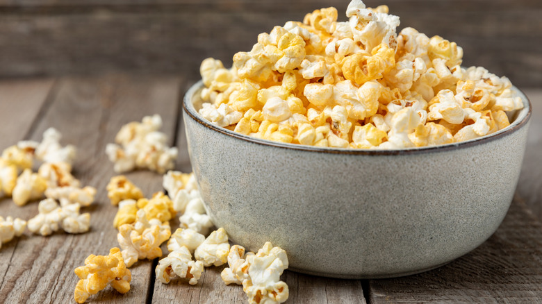 A bowl of buttered popcorn with a handful of kernels beside it on a wooden surface