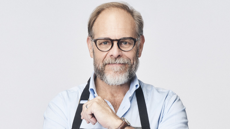 Alton Brown is smiling and wearing an apron over his dress shirt.