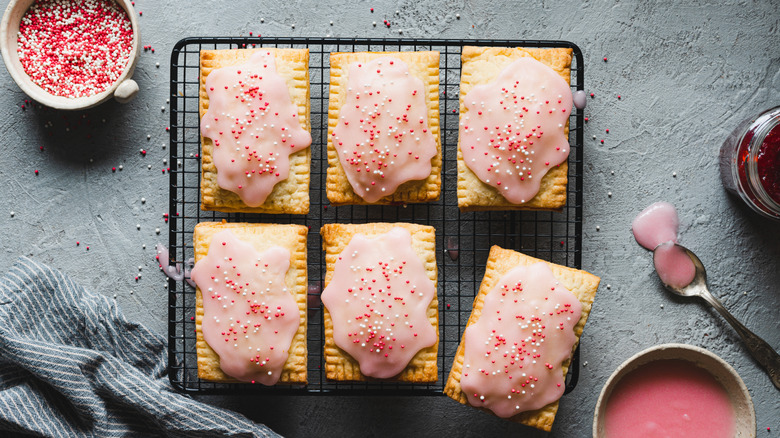 Homemade strawberry Pop-Tarts on a cooking rack near sprinkles, jam, and frosting, on a gray countertop.