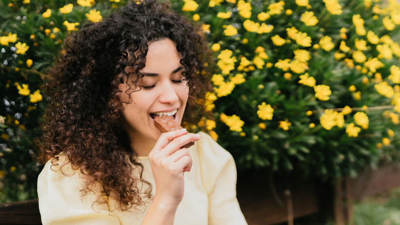 Curly-haired woman biting chocolate candy bar, surrounded by yellow flowers