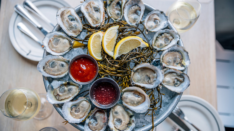 Fresh oysters on ice with lemon slices, dipping sauce, and seaweed on a dining table.