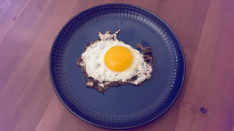 An egg fried in bacon grease on a blue plate, unbroken yolk sitting on a wooden table