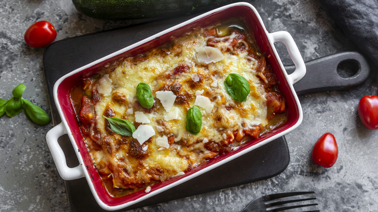 Lasagna with basil leaves and cheese in a white container with red interior. The container is sitting on a black cutting board which is in turn on a grey table with cherry tomatoes, basil and a single fork scattered about.