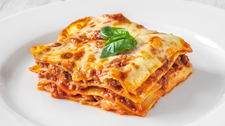 Square of four-layer lasagna with two basil leaves on top for decoration on a white plate