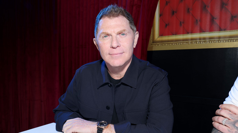 Bobby Flay sitting down at a table in front of a red curtain