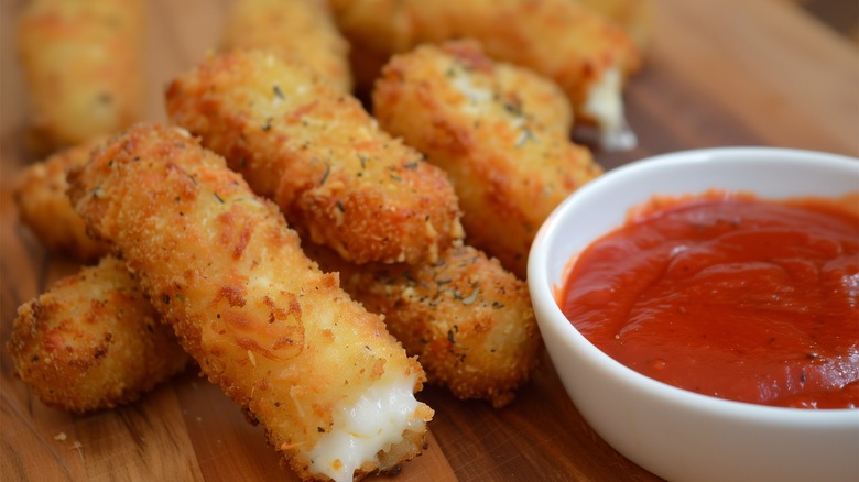 a pile of mozzarella sticks on a wooden surface near a small white bowl of marinara sauce