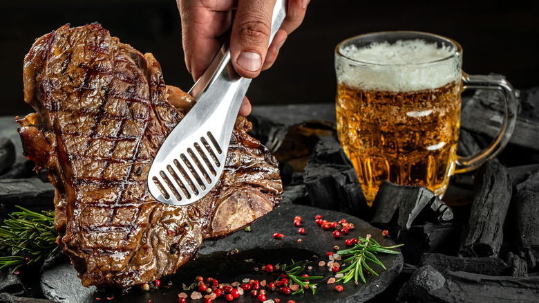 steak being placed with tongs on black plate glass and a mug of beer