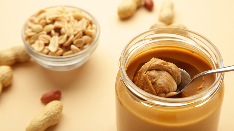 Open jar of peanut butter with a spoon and peanuts in the background