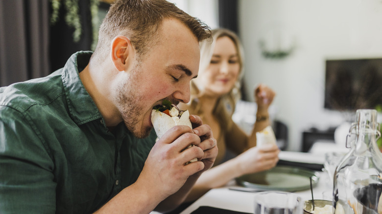 a man opening wide to take a big bite of a burrito while a woman looks on