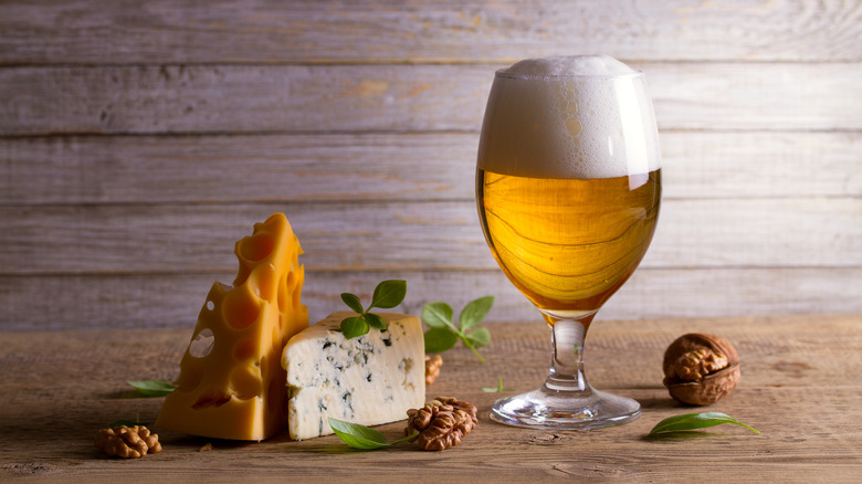 A glass of beer with cheese, walnuts, and green sprigs on a wooden background