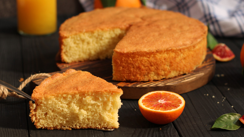 A slice of cake sits at the forefront of the image along with half an orange, behind these is the rest of the cake on a wiooden serving board, all of it sitting on a dark wooden surface.