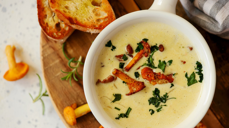 A bowl of creamy soup with parsley and mushrooms on top of a wooden cutting board with toast