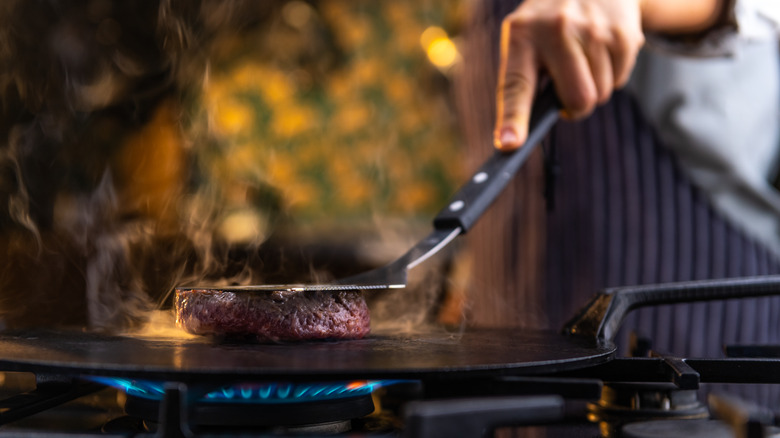 A cook searing a burger patty on a flat pan using a spatula