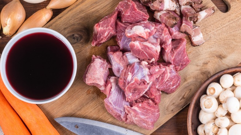 Wooden cutting board with prepared meat, vegetables, and bowl of red wine.