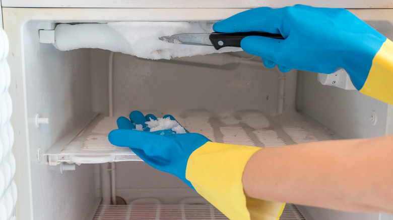 Someone wearing rubber blue gloves scraping ice off the inside of a freezer with a knife