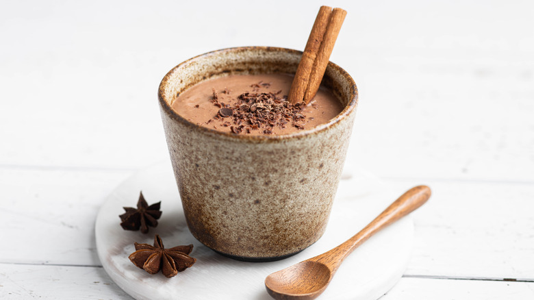 A cup of champurrado with a stick of cinnamon in it sits on a white plate