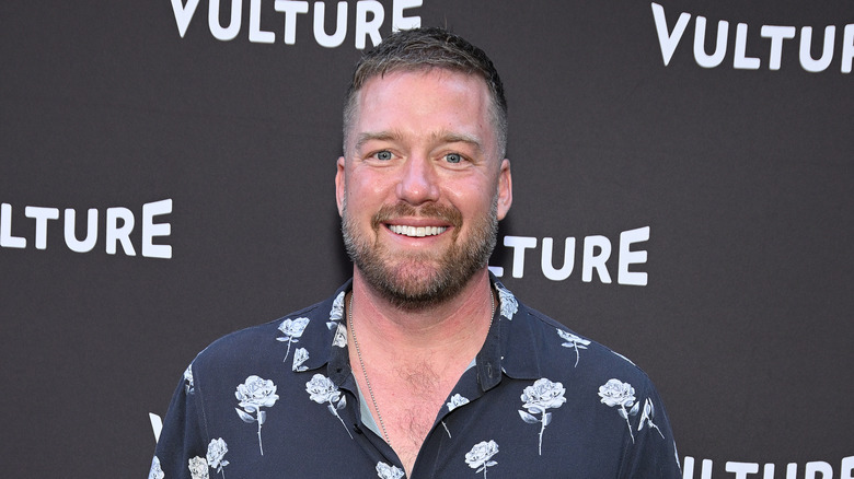 Tim Hollingsworth wearing a dark blue button down with white flowers, smiling in front of a step and repeat with the word 