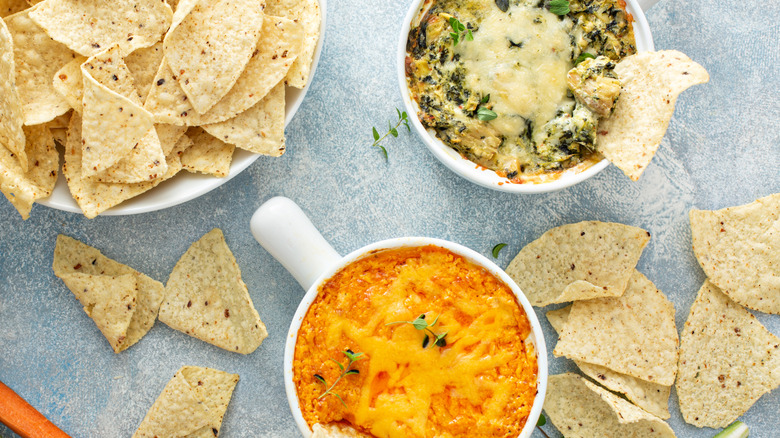 bowls of tortilla chips, spinach dip and buffalo chicken dip on a blue surface