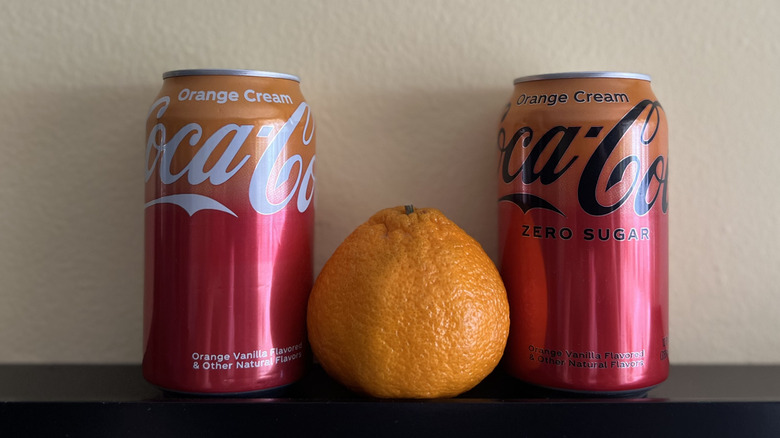 an orange sits between two cans of Coca-Cola Orange Cream