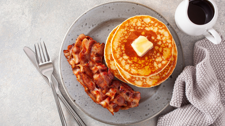 A plate wutg a stack of pancakes and strips of bacon on it beside a cup of syrup and silverware.