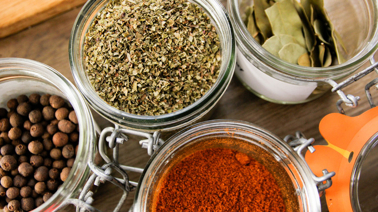 Jars of whole peppercorns, oregano, bay leaves, and a ground red spice