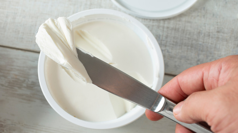 A person using a knife to scrape cream cheese out of a tub