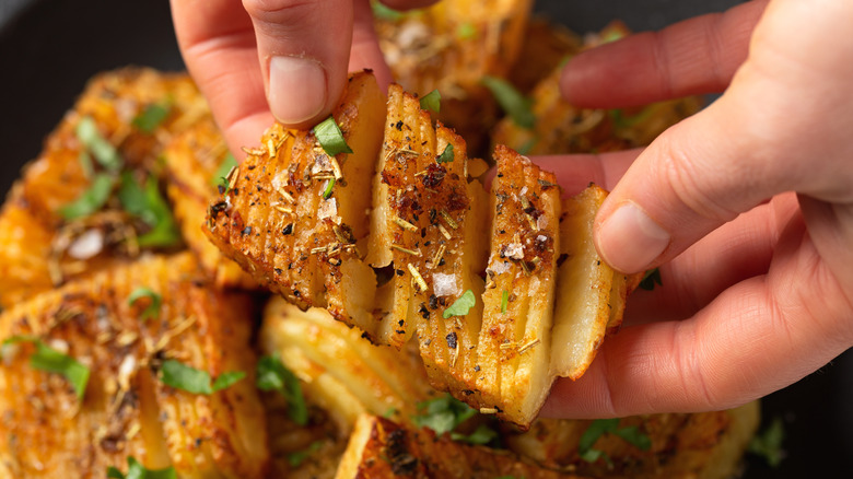 Someone pulls apart a cooked accordion potato, garnished with fresh herbs and spices, with more accordion potatoes in the background.