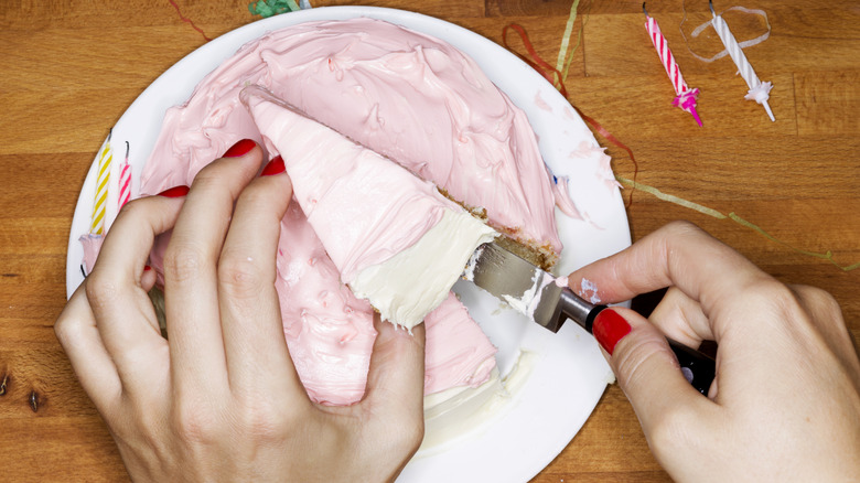 A hand with red nail polish cuts into a pink-frosted cake