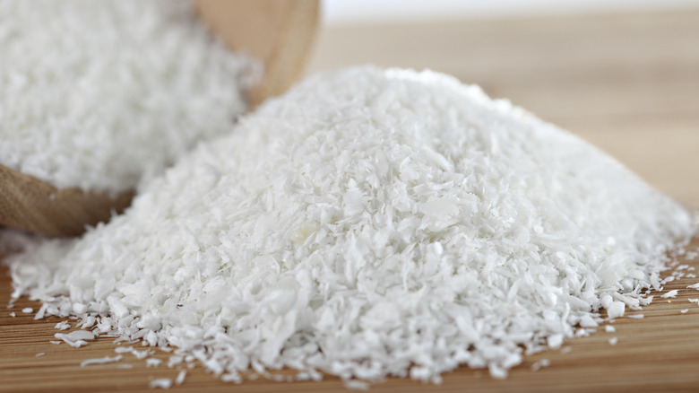 Pile of desiccated coconut spilling from a wooden bowl onto a wooden surface.